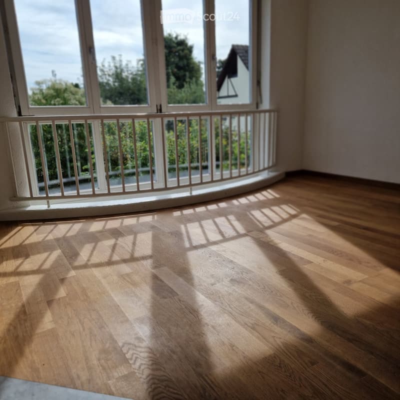 Empty room with wooden flooring and large windows overlooking a balcony area with a railing.