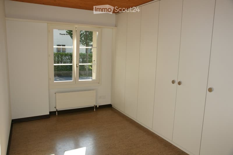 Empty room with white walls, wooden floor, built-in white wardrobes, and a window with a view of greenery outside.