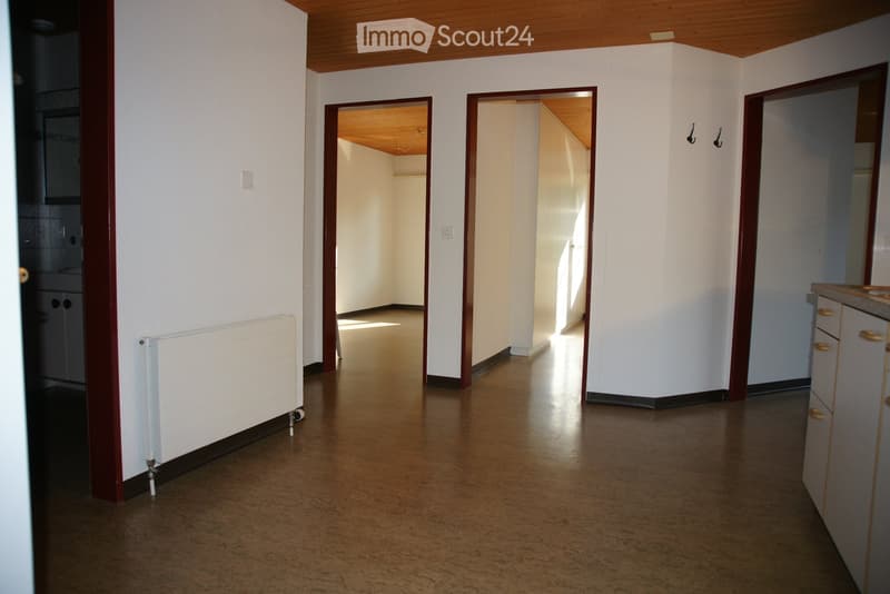 Empty hallway with wooden flooring, white walls, and several doors leading to other rooms. A radiator is visible on the left wall, and a small section of a kitchen is visible on the right.