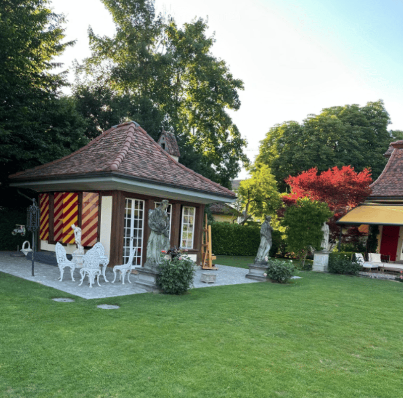 Small house, red roof, red and white painted walls, outdoor furniture, garden, statues, trees