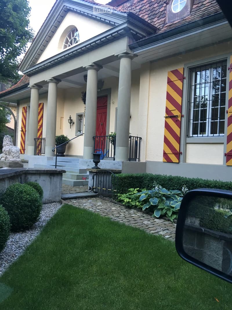 front view of two story house, beige exterior, red door, yellow and red shutters, front garden with lawn, bushes, lion statue, gravel pathway, black iron fence