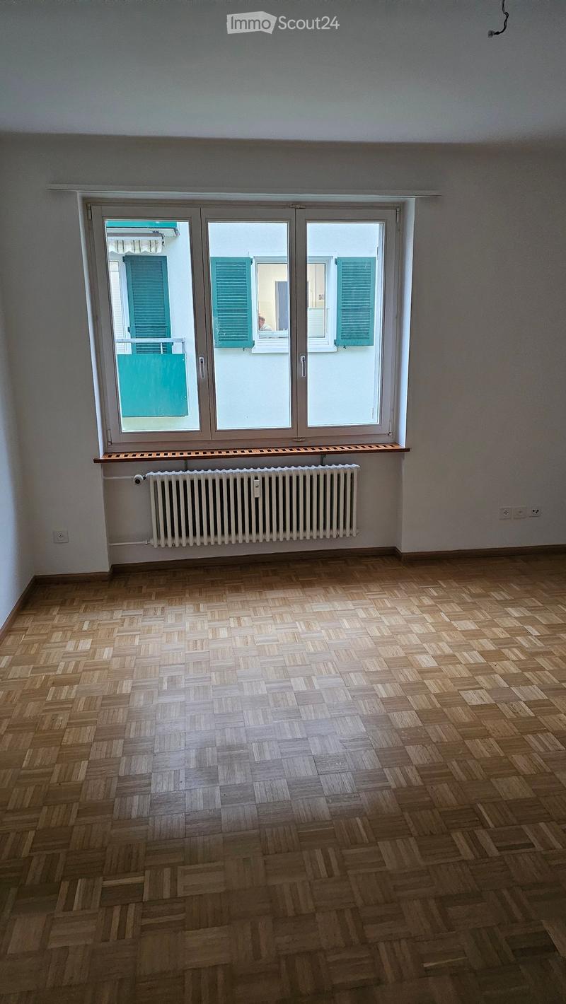 Empty room with wooden herringbone flooring, large window with shutters, radiator, and a small shelf above it.