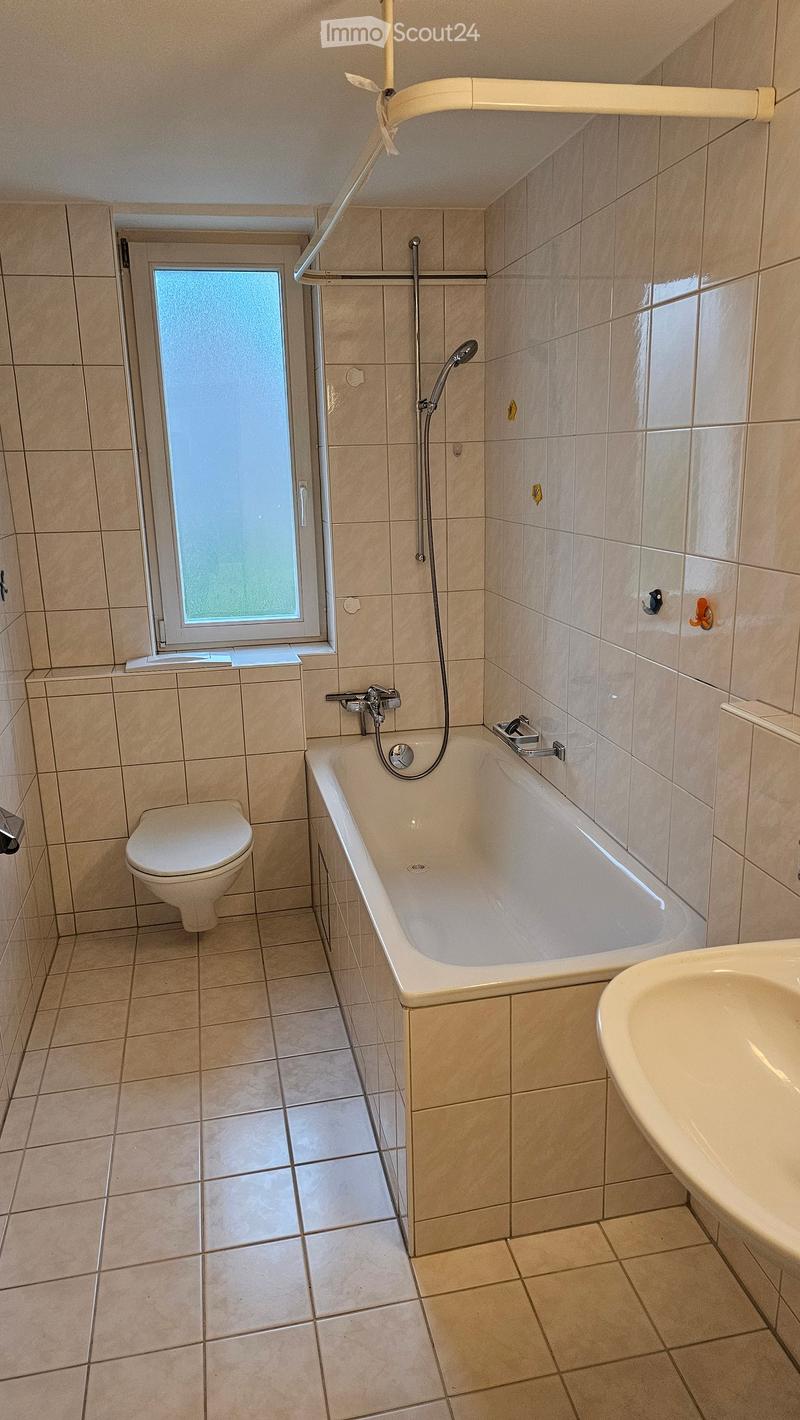 Bathroom with beige tiles, bathtub, toilet, sink, and showerhead. Features a frosted window and a safety grab bar.