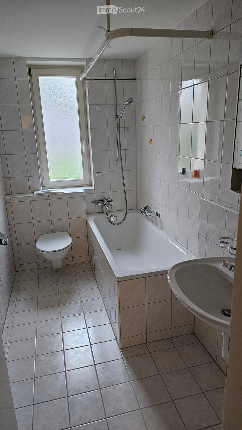 Modern bathroom with a bathtub, toilet, sink, and showerhead. The room features tiled walls and floor, a window with frosted glass, and a sleek, contemporary design.