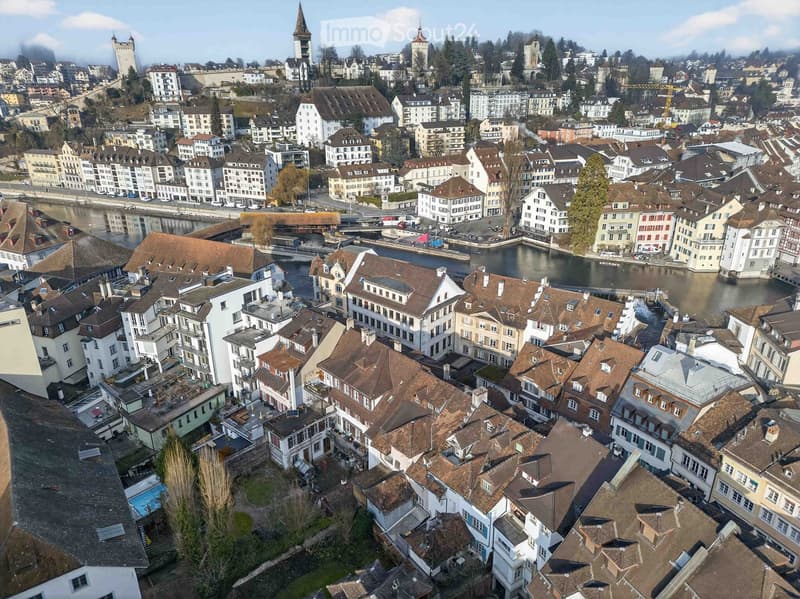 Aerial view of a European city with numerous buildings, a river, and a bridge. The architecture includes a mix of residential and commercial structures with varied roof styles, and there is a visible swimming pool in the foreground.