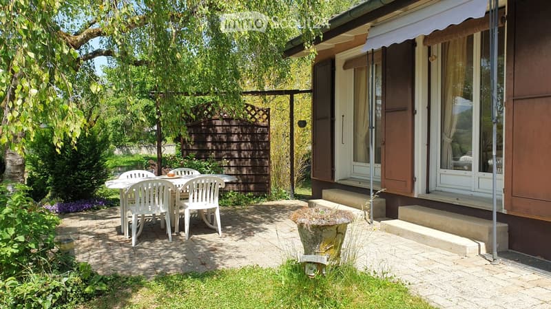 Garden area with a patio, white plastic chairs and table, steps leading to a house entrance, and lush greenery.