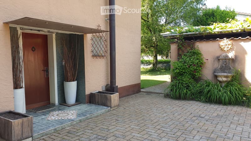 Entrance area of a house with a wooden door, decorative planters, a small fountain, and greenery. The pathway is paved with bricks, and the area is surrounded by trees and shrubs.