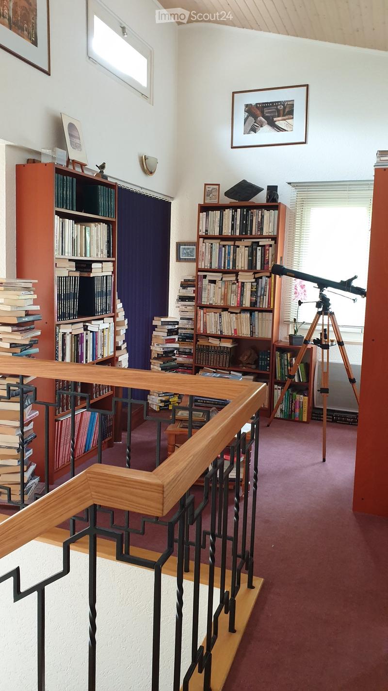 A cozy, well-lit room with bookshelves filled with books, a telescope on a tripod, and framed artwork on the walls. The room features a wooden railing and a purple carpeted floor.