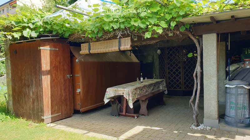 A covered outdoor seating area with a wooden table, bench, and wine bottle and glasses on the table. The area is partially covered by a wooden structure with vines growing on the roof.