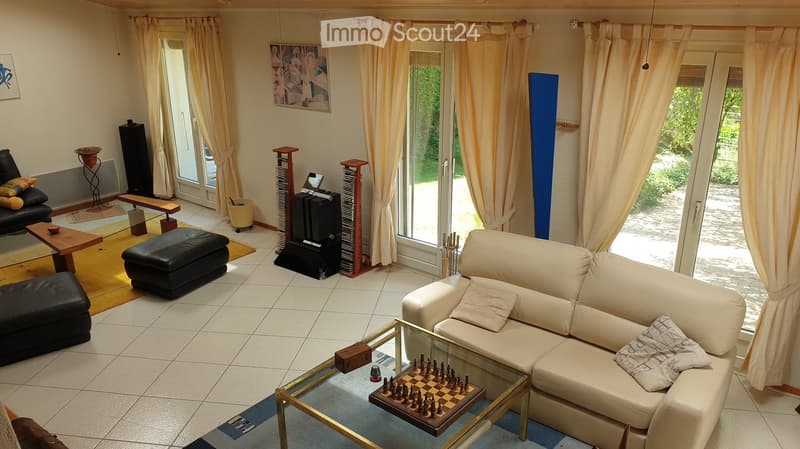 Bright living room with large windows and sliding glass doors leading to a garden. Features a cream-colored sofa, glass coffee table with chessboard, black ottoman, wooden side table, and a black leather chair. The room includes a speaker system, a lamp,