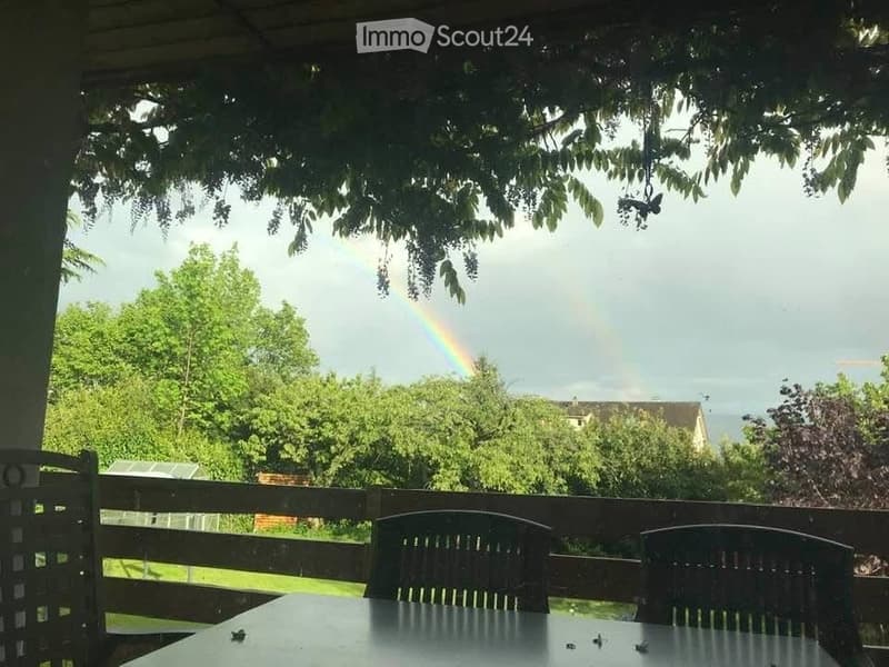 Outdoor patio with a table and chairs, surrounded by trees and a visible rainbow in the background.