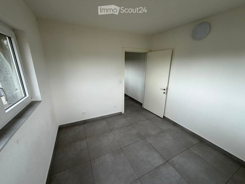 Empty room with grey tiled floor, white walls, and a large window with a view. No furniture or equipment present.