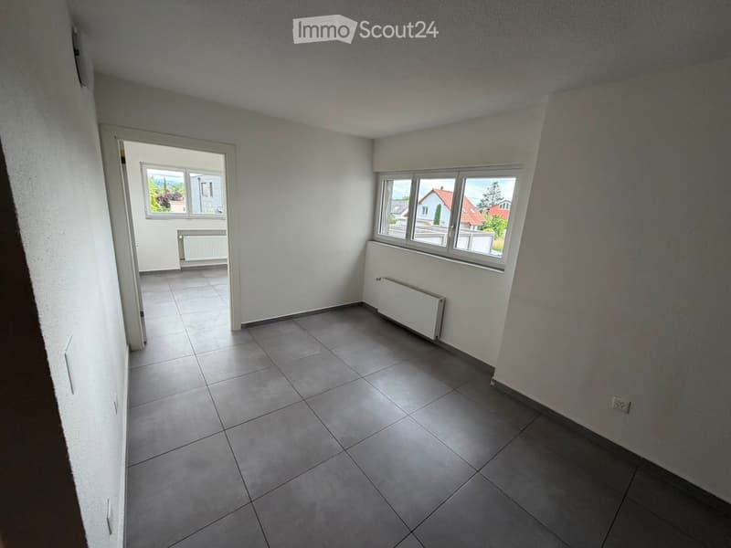 Empty room with tiled floor, white walls, and two windows offering a view of the outside. Radiators are visible under the windows.