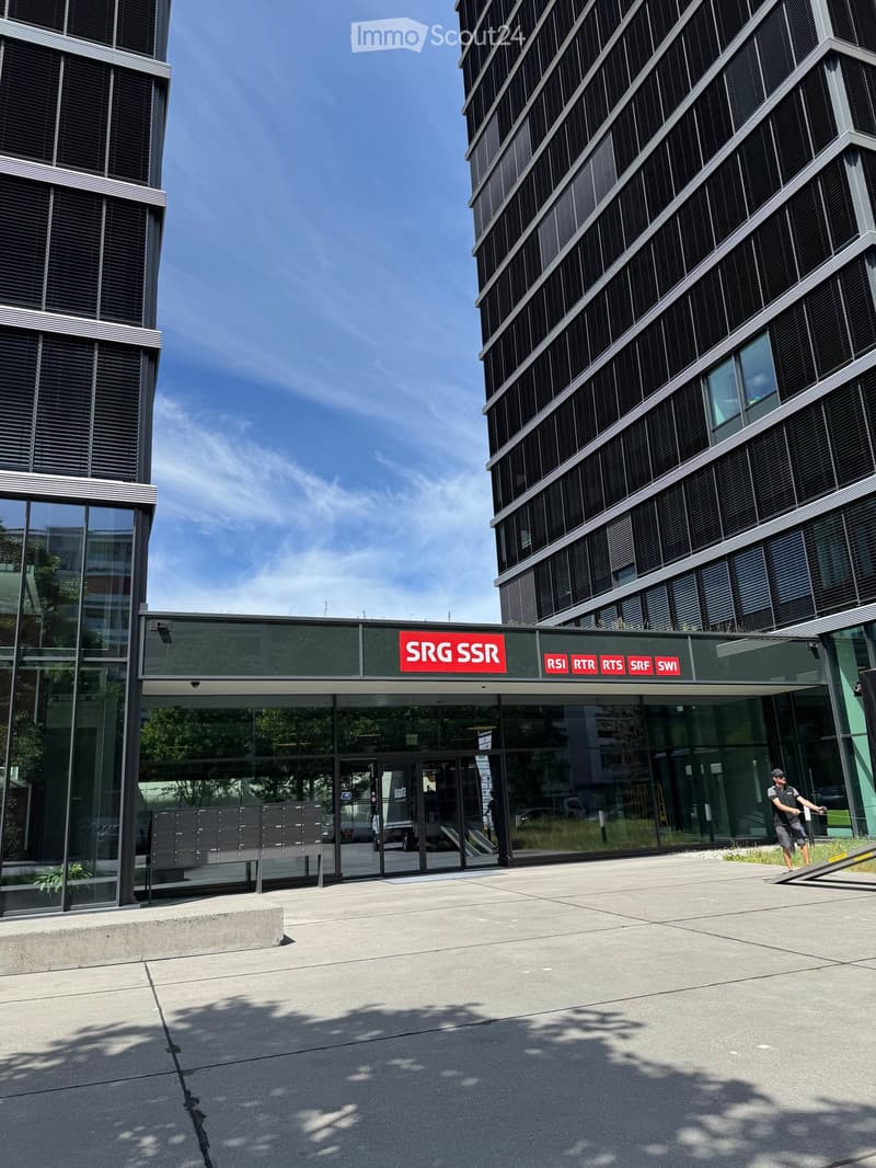 Modern building entrance with glass doors and a sign reading "SRG SSR" and other logos, surrounded by contemporary architecture and a clear blue sky.