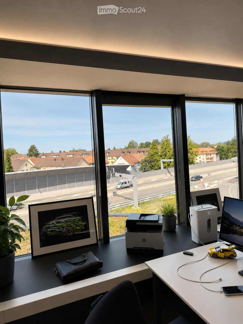 Modern office space with large windows overlooking a cityscape, featuring a desk with a printer, screen, laptop, cellphone, picture frame, plant, and a small toy car.