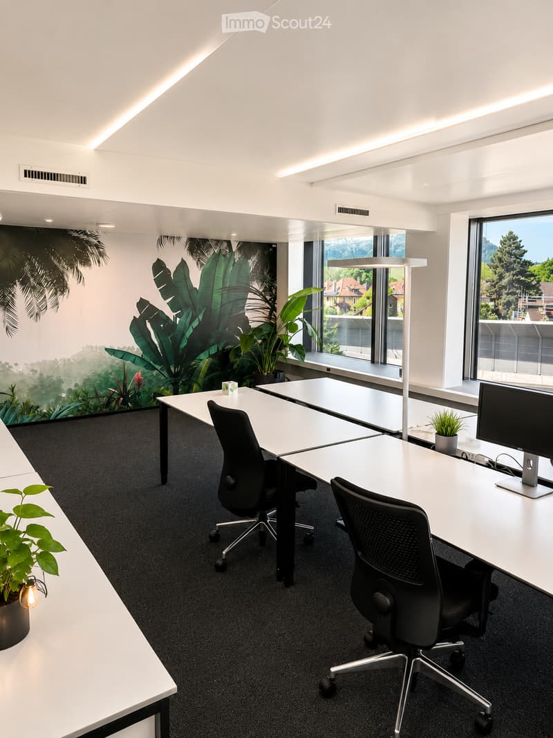 Modern office space with white tables, black office chairs, a computer monitor, potted plants, and a decorative wall mural.