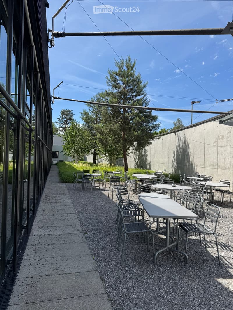 Outdoor seating area with tables and chairs arranged on gravel, adjacent to a modern building with large glass windows and a clear blue sky.