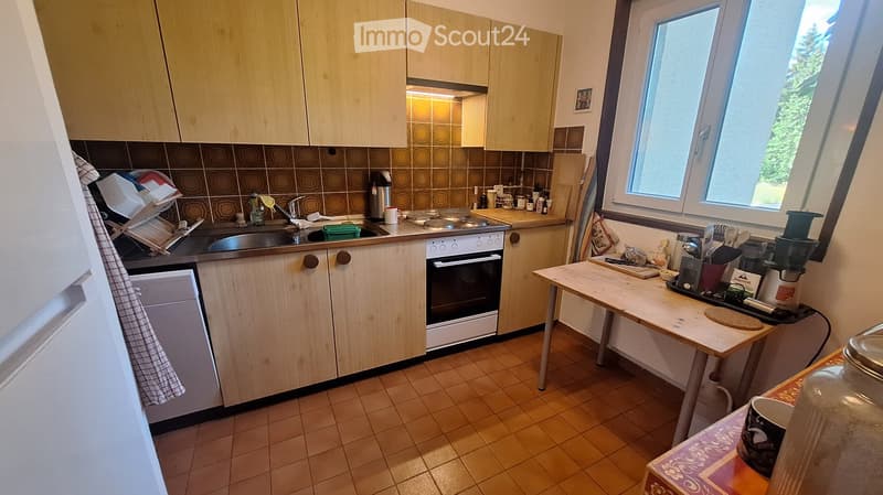 A well-lit kitchen with light wooden cabinets, a tiled backsplash, and various appliances including a sink, oven, and refrigerator. A small table with a coffee maker and other kitchen items is visible near the window.