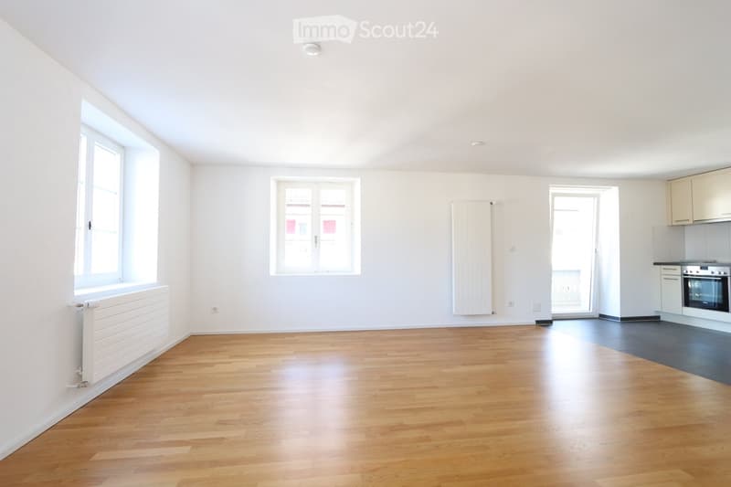Empty room with wooden flooring, white walls, and large windows. Radiators are visible on the walls, and a small kitchen area is visible in the background.