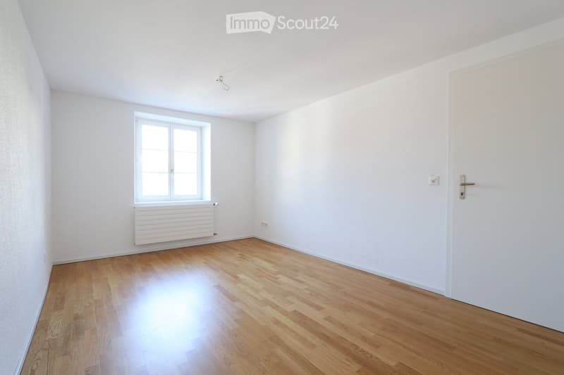 Empty room with white walls, wooden flooring, a window with white frame, and a radiator below it. No furniture or equipment present.