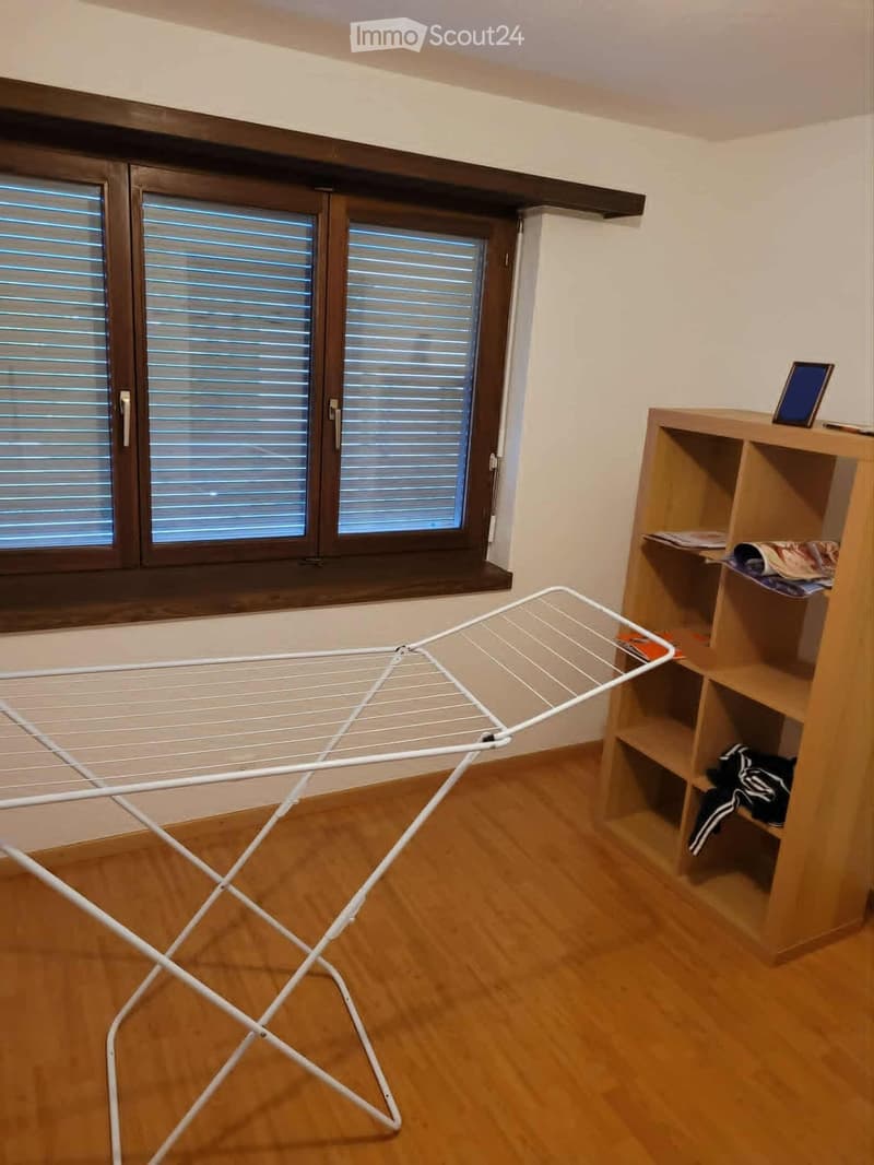 A small, unfurnished room with wooden flooring, large windows with blinds, a wooden shelf holding a screen and some folded clothes, and a white drying rack.