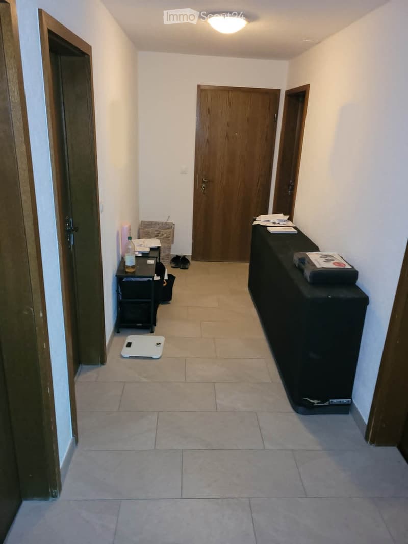A narrow hallway with tiled flooring, white walls, and wooden doors. There is a small table with a bottle and papers, a black cabinet with items on top, and a white scale on the floor.