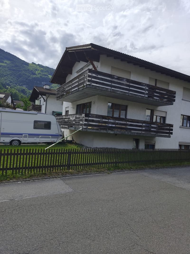 Two-story house with a balcony, surrounded by a wooden fence and a trailer parked nearby.