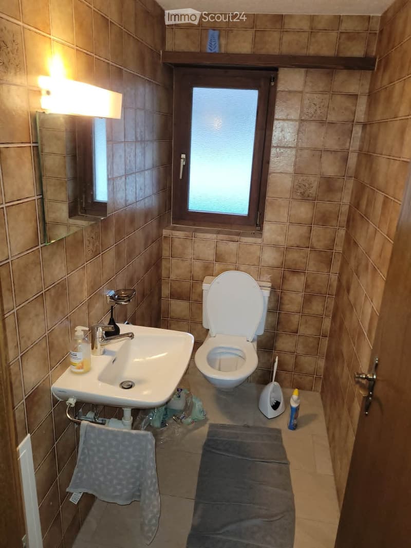 A small bathroom with beige tiled walls and floor, featuring a white sink with a chrome faucet, a toilet, a urinal, a towel, and a bottle on the sink counter.