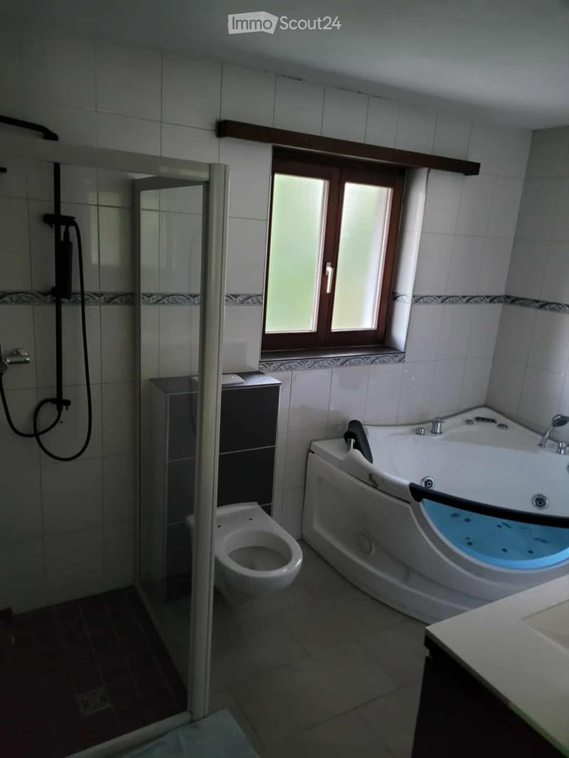 Bathroom with a bathtub, toilet, shower, and sink. The room features tiled walls and a window with wooden frames.