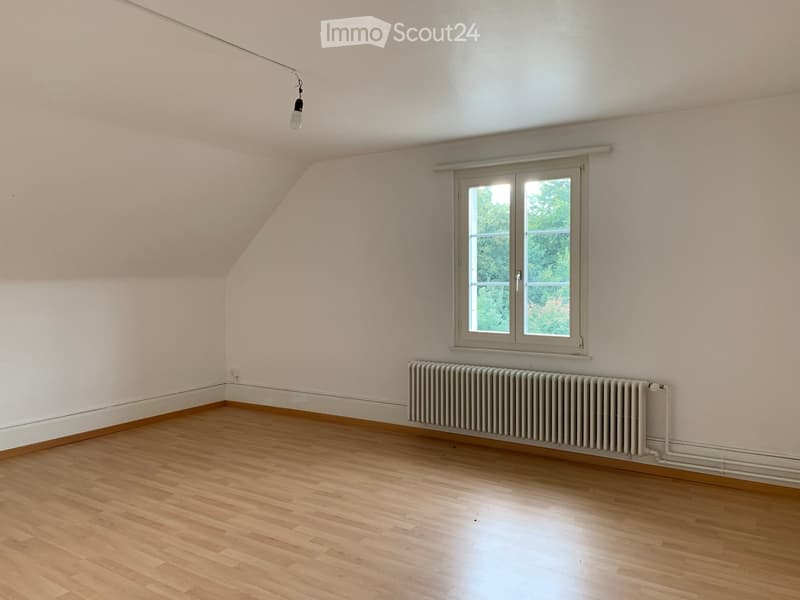 Empty attic room with wooden flooring, white walls, a single window, and a radiator. The ceiling has a sloped design typical of attic spaces.