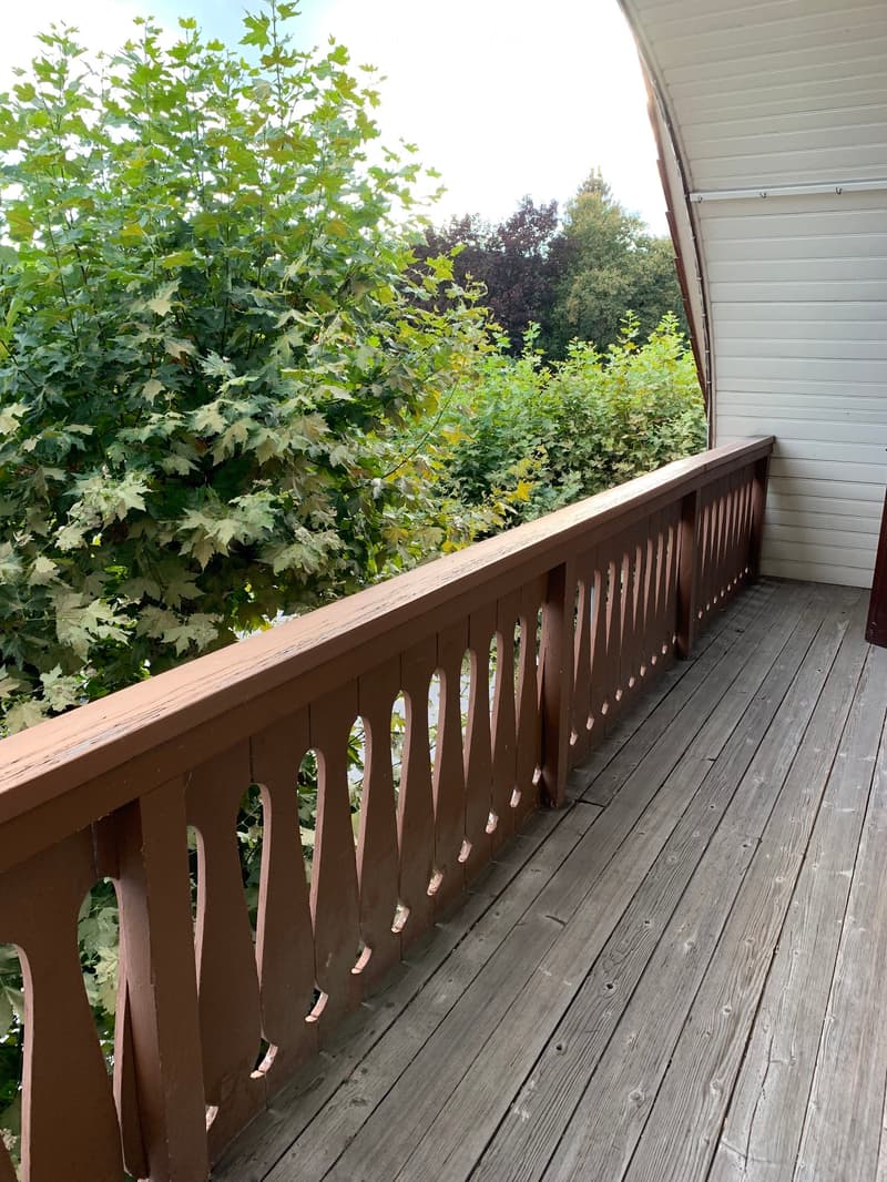 Wooden deck with a brown railing featuring decorative balusters, overlooking a lush green garden area.