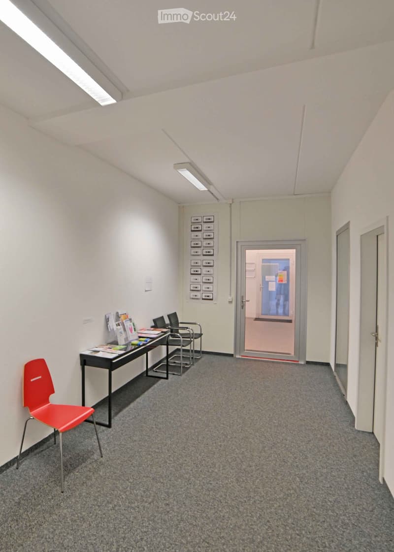 A small, well-lit room with a grey carpeted floor, white walls, and a slanted ceiling. It features a black table with brochures and a laptop, a red chair, and several black chairs arranged near the table. A door with a glass panel leads to another area.