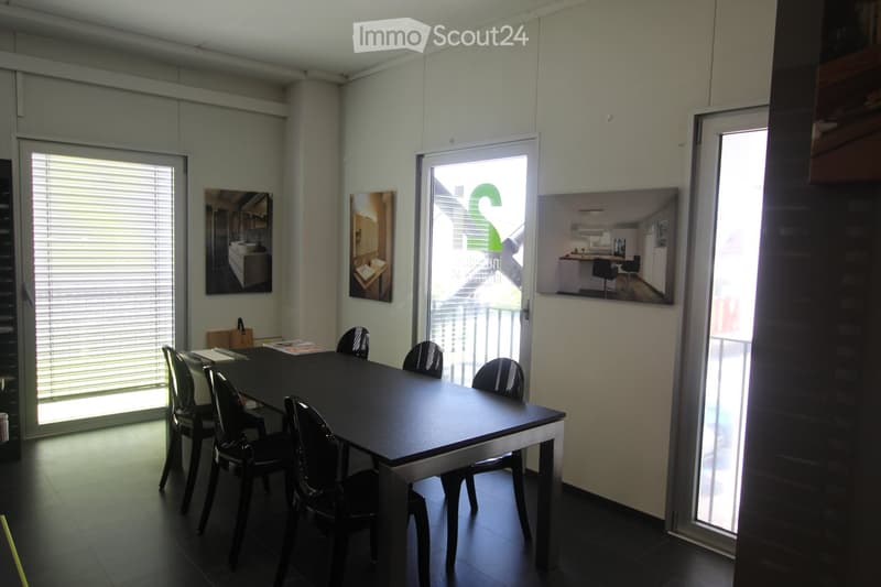 Bright room with a black table and six black chairs, large windows with blinds, and several picture frames on the walls.