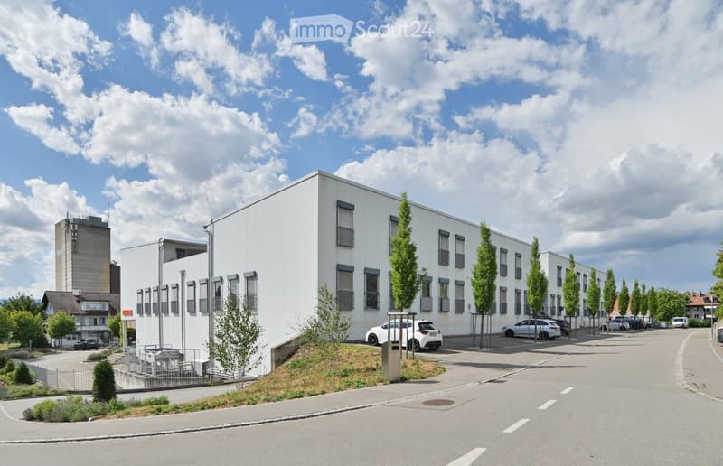 Modern white building with multiple windows and a raised ground floor, surrounded by trees and parked cars.