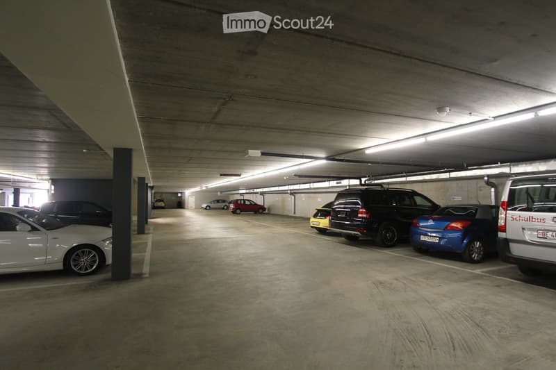Underground parking garage with concrete floors and walls, 6 cars parked, entrance in the distance, white ceiling with fluorescent lighting