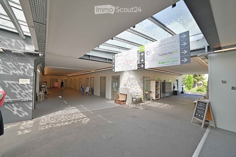 Modern, spacious hallway with a high ceiling and large windows allowing natural light. Directional signs point to various rooms and areas, including "Bewegung & Entspannung" and "Ergotherapie." The floor is concrete, and there are a few small tables and c