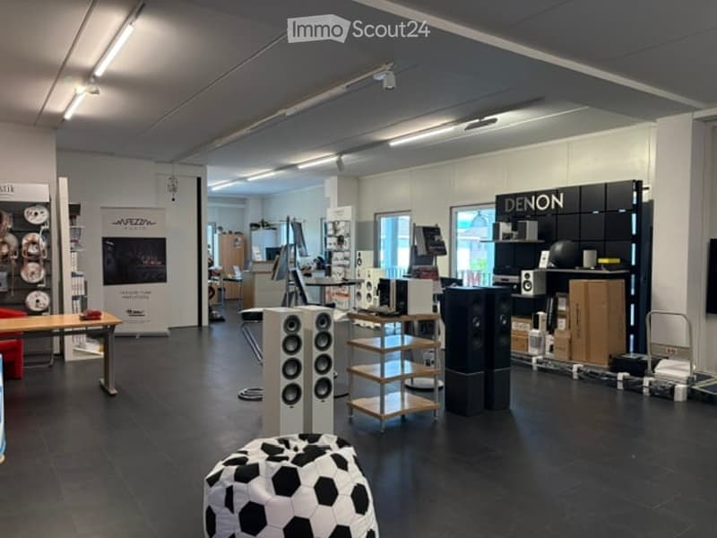 A modern, well-lit showroom with a variety of audio equipment displayed on shelves and stands. A black and white soccer ball stool is placed in the foreground, and a table with a red chair is visible on the left side.