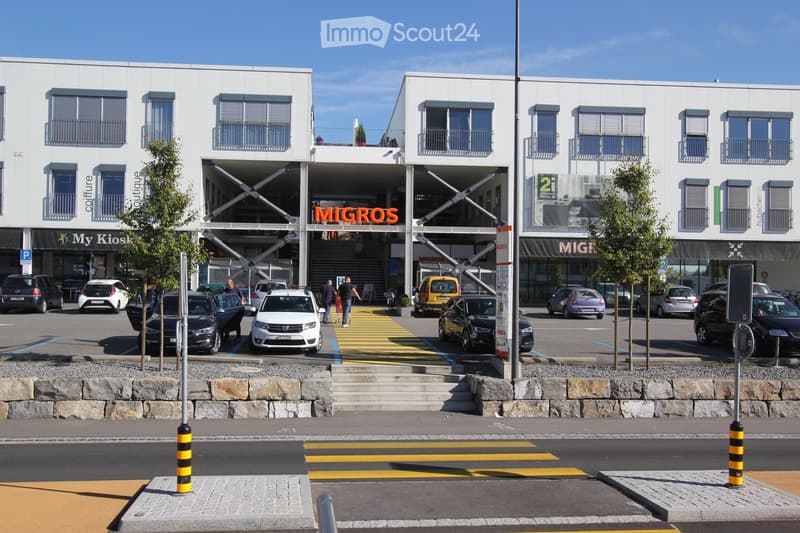 Modern commercial building with several floors, multiple windows, parking lot with several cars parked, trees and bollards, sign reading 'Migros'.
