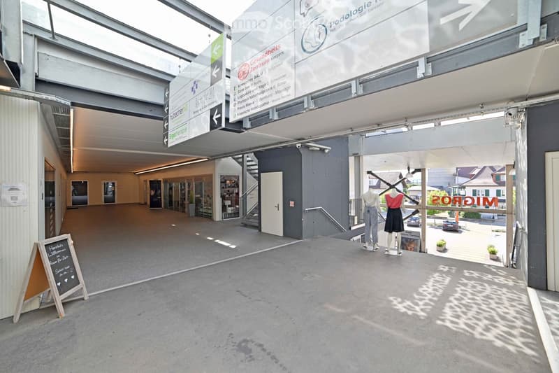 Modern building entrance with signage for various health services, including podology, physiotherapy, and massage. The area features a spacious, open lobby with glass walls, a staircase, and a small display area with mannequins.