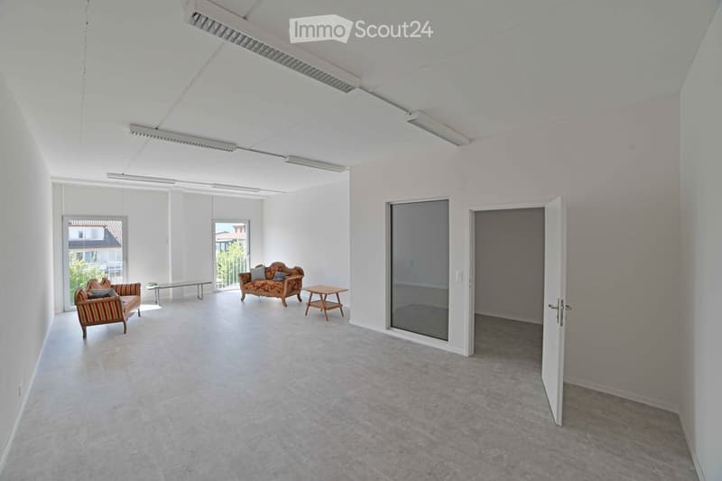 Bright, open room with white walls and large windows, featuring a patterned sofa, a small table, and a chair.