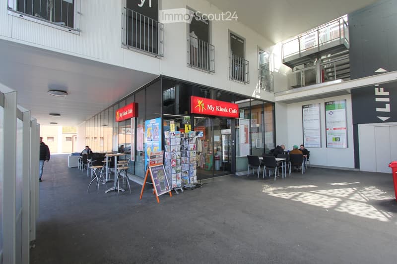 Modern building with multiple floors, storefronts, outdoor seating area, and a trash bin visible.
