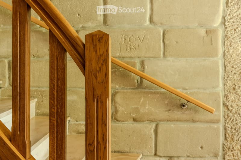 Close-up of a wooden staircase railing with a polished finish, set against a beige brick wall with an engraved date and initials.