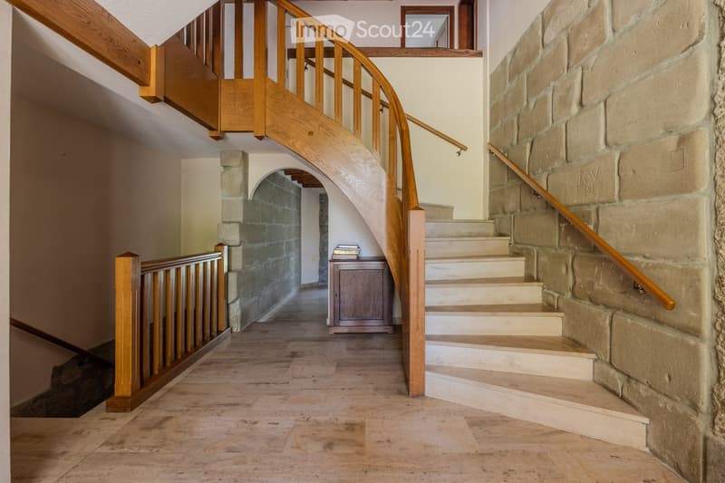 Spacious hallway with wooden flooring, stone walls, and a curved wooden staircase leading upwards. A small wooden cabinet and books are visible in the background.