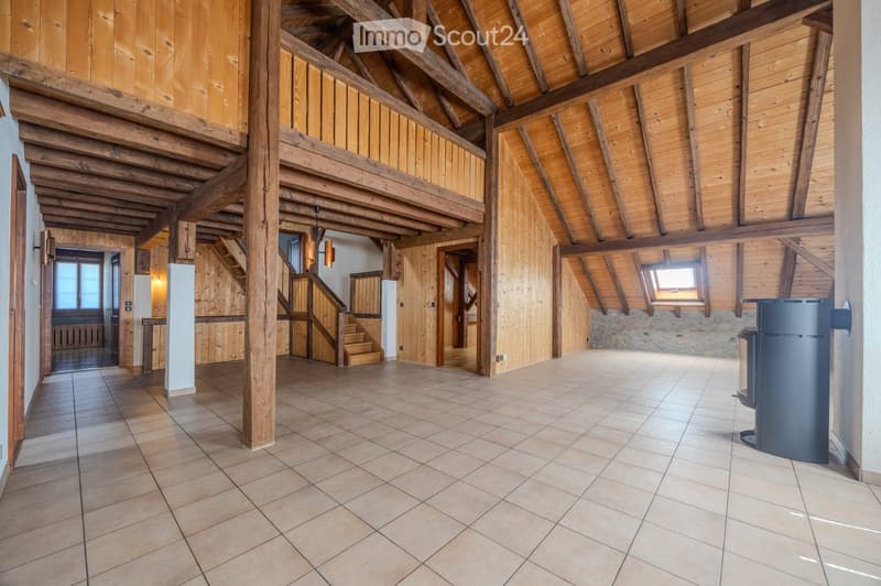 Spacious hallway with exposed wooden beams and a high, vaulted ceiling. Features tiled flooring, wooden staircase leading to an upper level, and a small seating area with a table and chairs.
