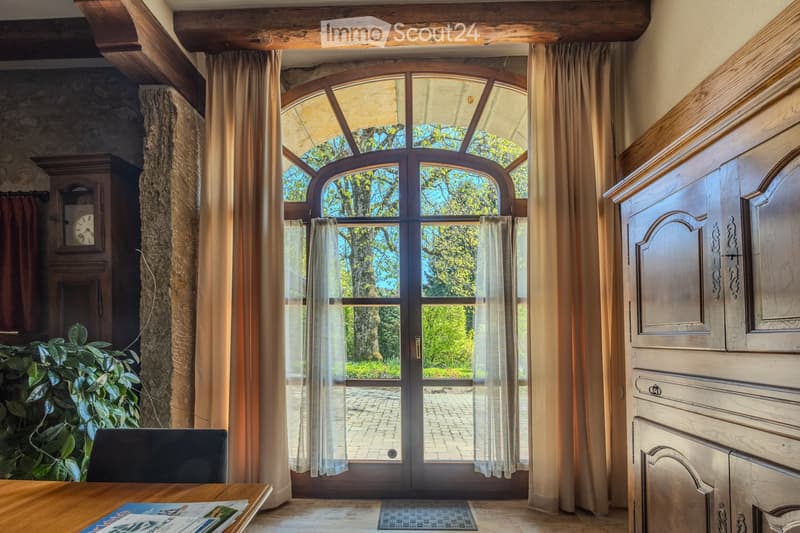 Elegant room with wooden ceiling beams, large arched windows with curtains, a wooden table, a chair, a cabinet, and a clock on the wall.