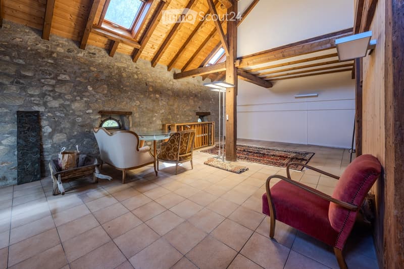 A cozy living room with exposed wooden beams, stone walls, and a fireplace. The room features a sofa, chairs, a glass coffee table, and a floor lamp.