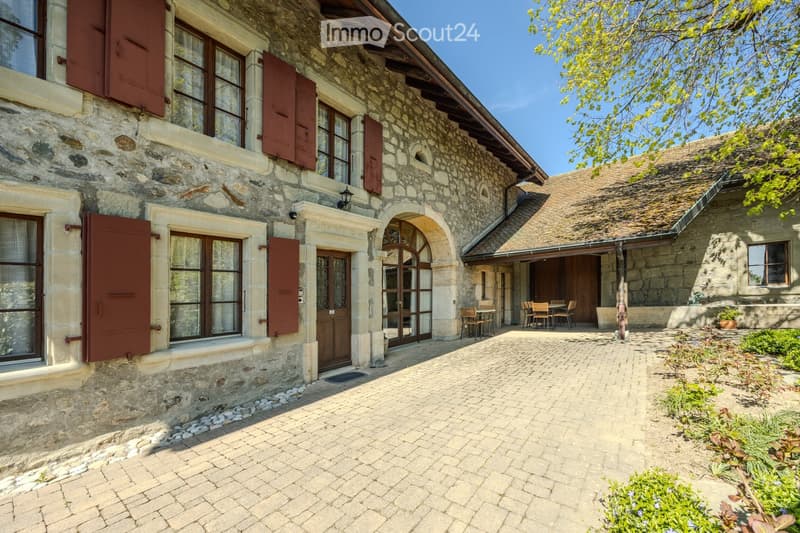 A stone house with red shutters, a wooden door, and a covered patio area with outdoor furniture. The house features a tiled roof and is surrounded by a cobblestone courtyard with greenery.