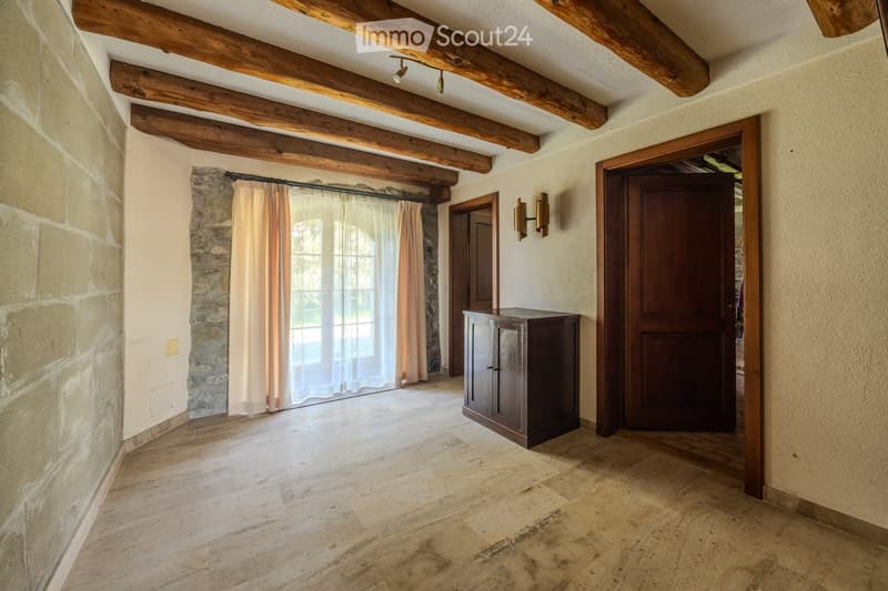Spacious hallway with exposed wooden beams, stone walls, wooden floor, large window with curtains, small wooden cabinet, and two doors.