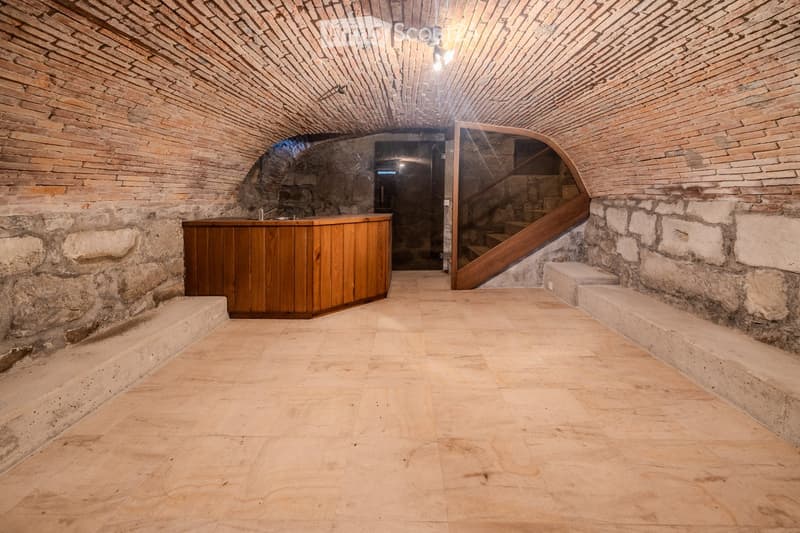 A rustic, vaulted room with exposed brick walls and a wooden bar counter. The space features stone flooring and a staircase leading upwards.