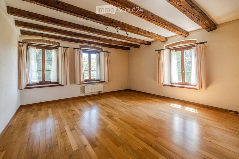 Empty room with wooden floors, exposed wooden beams, large windows with curtains, and a radiator on one wall.